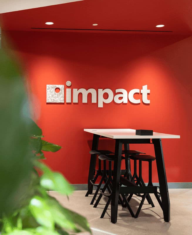 Common area with bar table and stools, Impact Networking logo on the red wall behind it | Impact Networking office in Riverside
