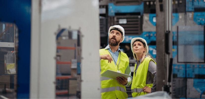 Two manufacturing workers in hard hats and high visibility yellow vests in a warehouse