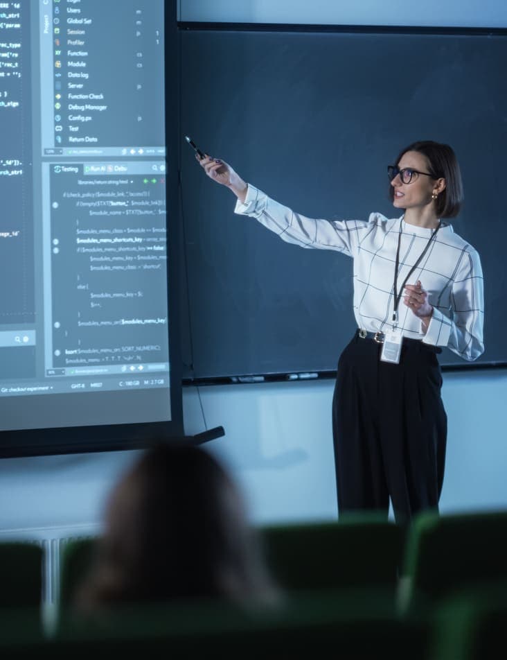 A teacher in front of a classroom pointing to coding data on a projection screen