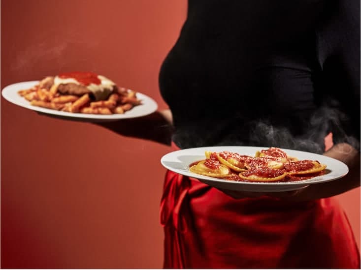 A server holding two steaming plates of saucy pasta dishes from Beggars Pizza.