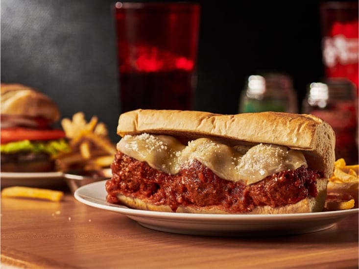 A large meatball sub topped with cheese sits on a plate with various Beggars Pizza menu items in the background.