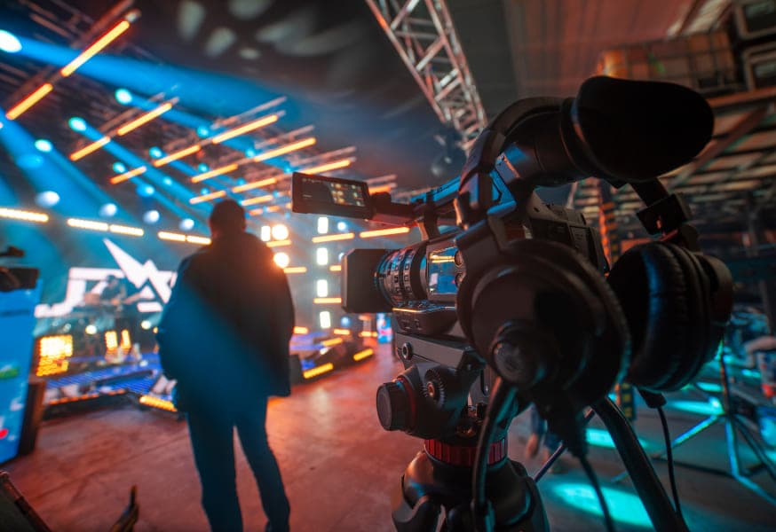 A video camera pointed at a person standing in front of a colorfully-lit studio set