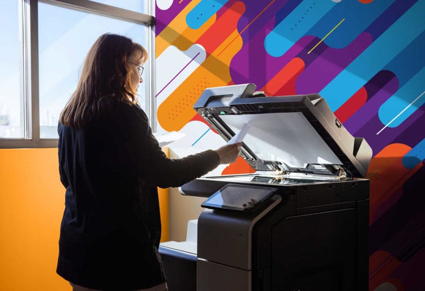 Woman standing at a office printer checking a document with a window to the left and a colorful wall behind the printer